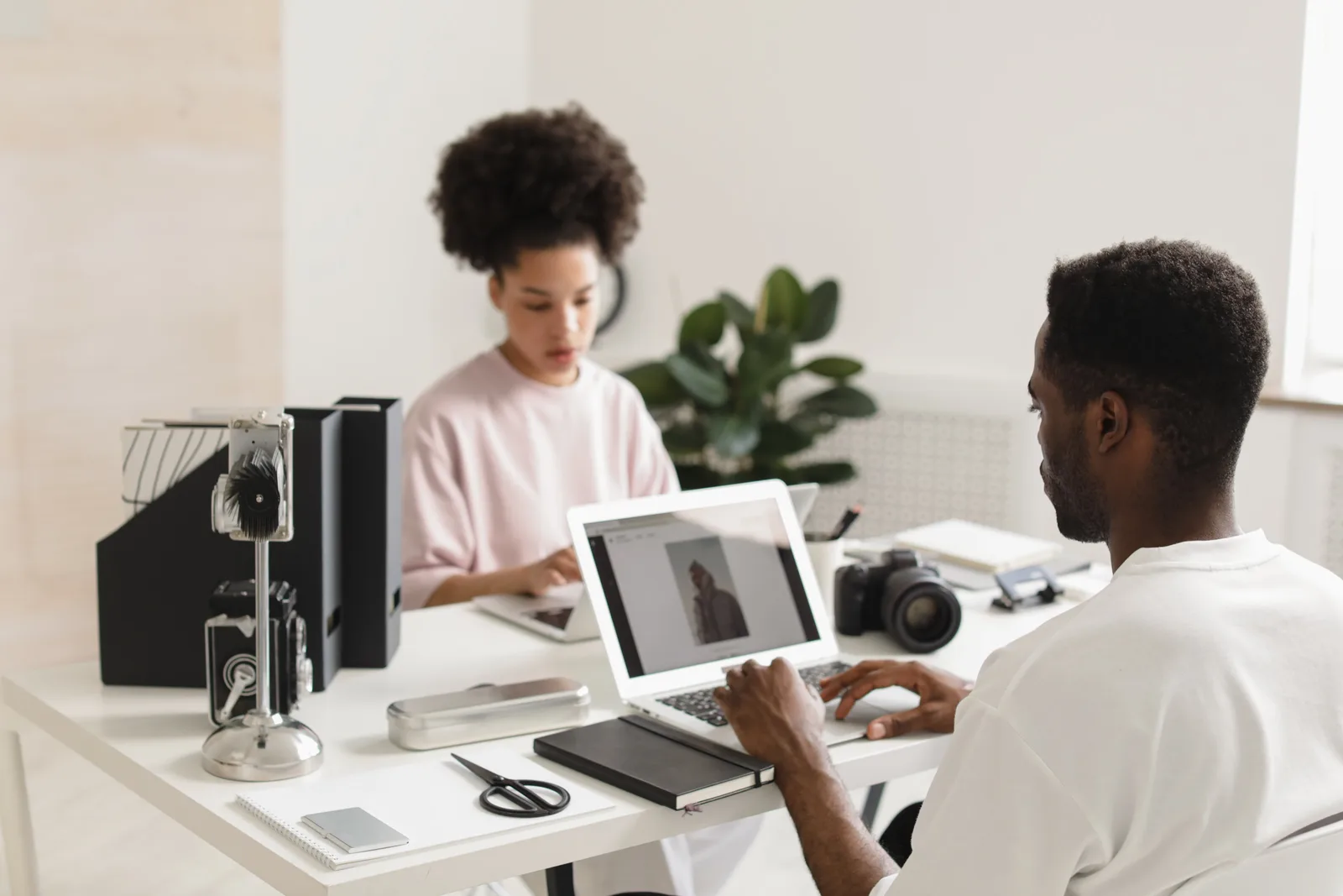 Two creative professionals collaborating in a bright, modern office setting with laptops and camera equipment.