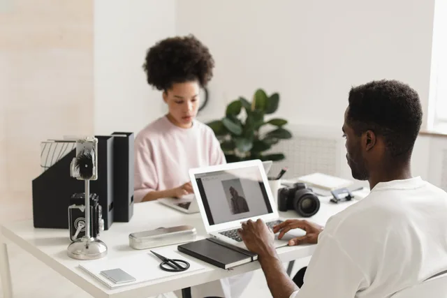 Two creative professionals collaborating in a bright, modern office setting with laptops and camera equipment. Creator brand ownership and partnerships